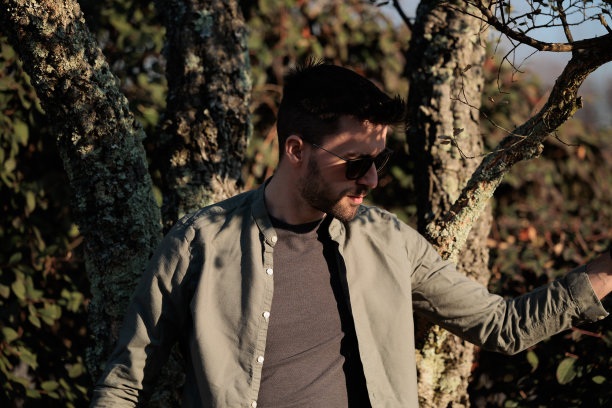 照片中，一位年轻的意大利成年男子挽着腰望向别处，阳光照在他身上，他穿着宽松轻便的长衣服，戴着一副太阳镜，在午后靠着一棵树站着图片下载