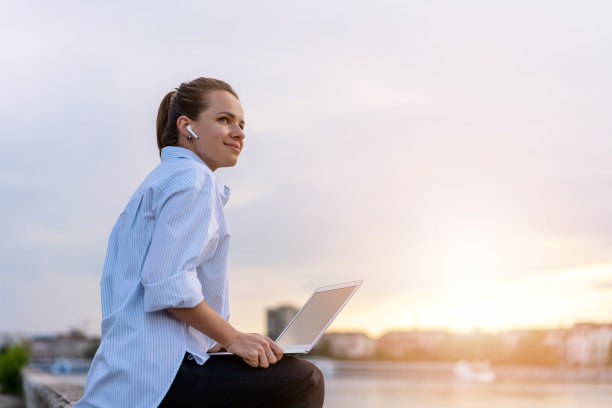 一位年轻的城市女性在办公室工作之外，拿着笔记本电脑坐在海滨欣赏日落。图片下载
