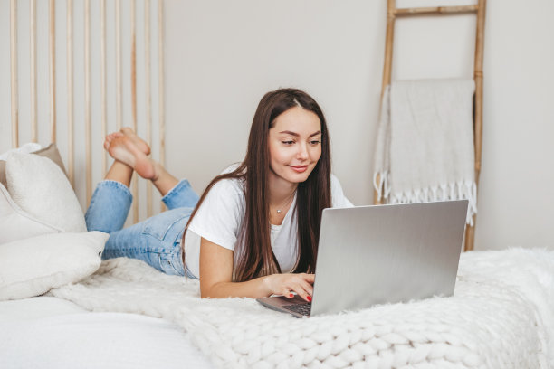 一名年轻女子趴在床上，一边用笔记本电脑工作，一边上在线英语课程图片下载