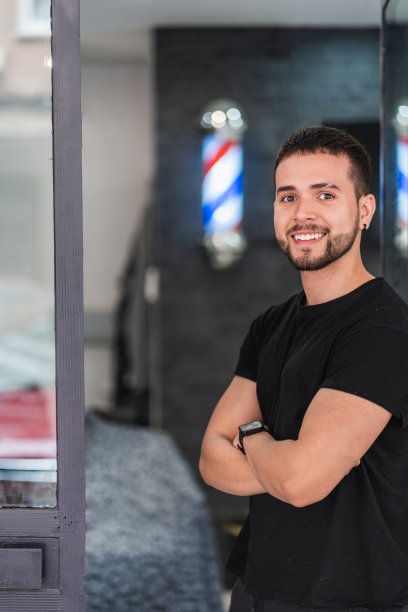 理发师双臂交叉倚靠在理发店门上的半长垂直肖像。图片下载