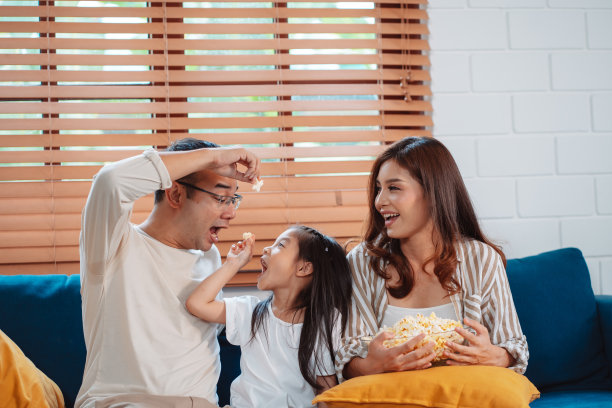 亚洲家庭由父母和女儿在家里客厅的沙发上看电视或电影，吃爆米花组成。享受放松的快乐。图片下载