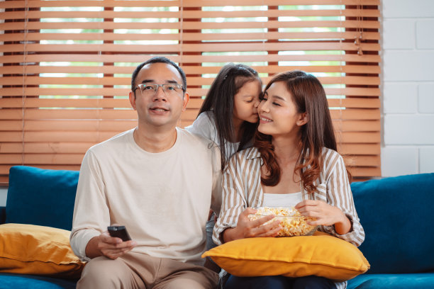 亚洲家庭由父母和女儿在家里客厅的沙发上看电视或电影，吃爆米花组成。享受放松的快乐。图片下载