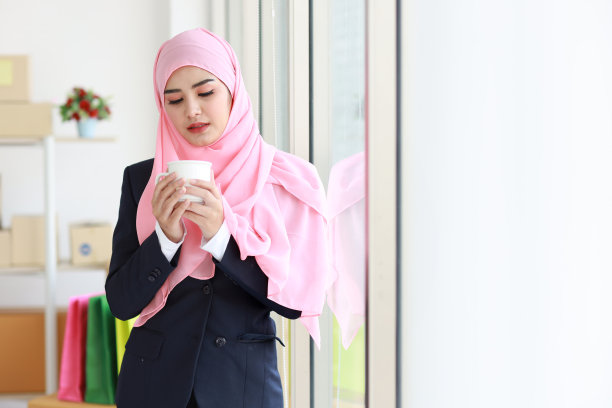 身着穆斯林蓝色西装的亚洲宗教妇女喝着咖啡，微笑着，头上戴着粉红色的围巾，背景是包裹盒。创业小公司中小企业自由职业女孩在家工作，笑脸相迎图片下载