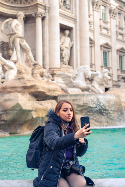 在意大利罗马度假期间，年轻的拉丁女游客穿着保暖的衣服，背着背包和专业相机，坐在特雷维喷泉边，用智能手机自拍图片下载