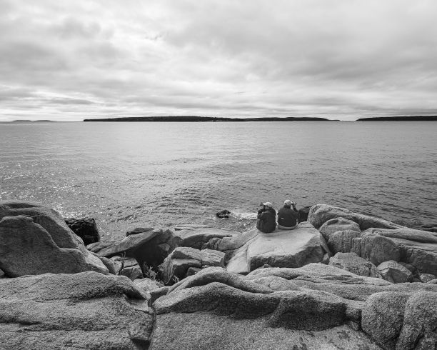 两个人坐在岩石海岸线上的灰度照片，面对着平静的水体。图片下载