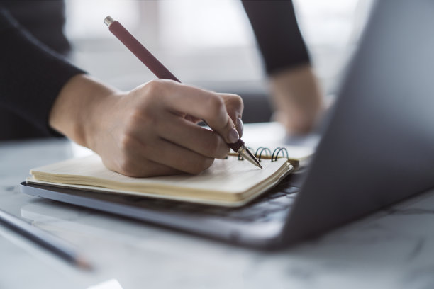 这张照片近距离拍摄了一位女士的手，她在一台别致的笔记本电脑上的记事本上写字，背景没有对焦图片下载