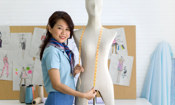 时装设计师室。年轻的亚洲裁缝在条纹围巾使用卷尺，测量人体模型的大小。布料的图案在软木板上。图片下载