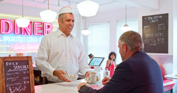 身穿白色衬衫和纸帽的男人在20世纪50年代风格的餐厅里与成熟的顾客交谈图片下载