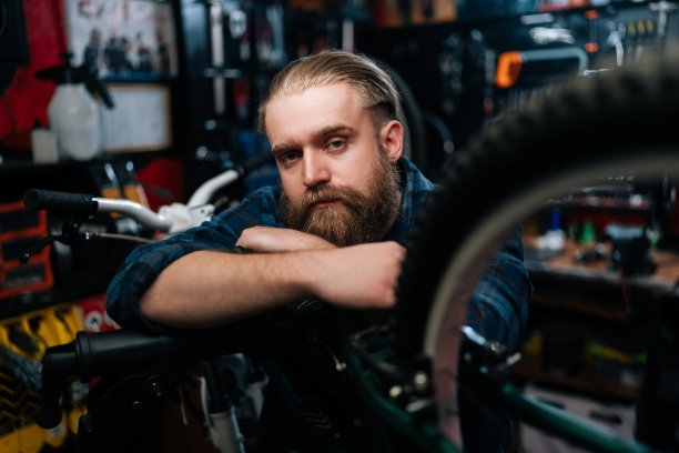 自行车修理工站在自行车上的特写肖像，在黑暗的内部修理自行车车间，严肃地看着相机。图片下载