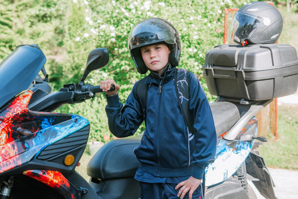 年轻可爱的少年戴着摩托车头盔，背景是摩托车。儿童安全。头上戴着头盔的男孩图片下载