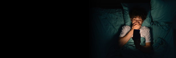 深夜，一个被吓坏的年轻人躺在床上，在黑暗的卧室里用手机看恐怖电影图片下载