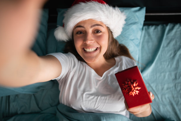 积极的女孩抱着礼盒躺在家里的床上自拍。圣诞节的早晨图片下载