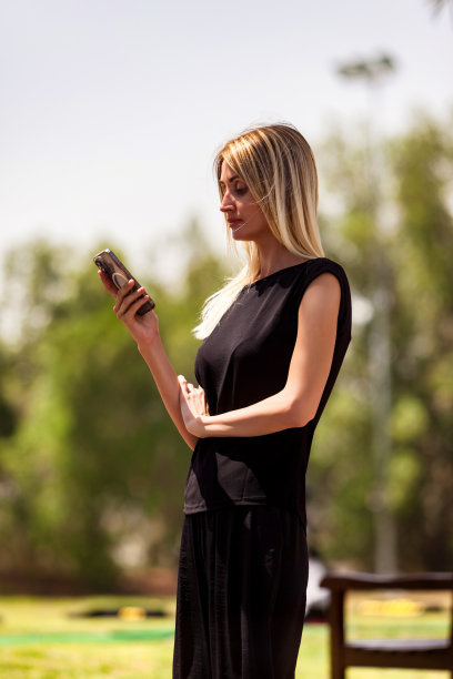 漂亮的年轻时尚的女商人的肖像，穿着黑色的衣服，手里拿着手机在城市公园。金发商人在大自然中使用手机。业务活动概念。复制广告文本空间图片下载