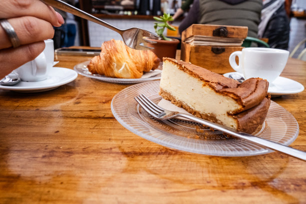 早餐。女人手里拿着叉子拿着一块芝士蛋糕放在一个玻璃板上，桌子上有一杯咖啡放在一个用金属勺子的盘子上，玻璃板上放着一个牛角面包和一杯茶。一切都在一张天然木桌上。美食。加那利群岛的特内里费岛图片下载