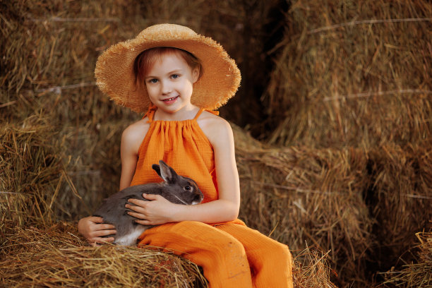 可爱的小女孩穿着橙色的裙子，戴着草帽，和一只棕色的兔子坐在稻草捆上图片下载