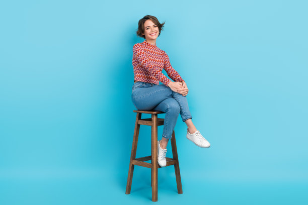 照片可爱自信的女士坐在高酒吧椅子点饮料酒保穿衬衫裤子运动鞋孤立的蓝色背景图片下载