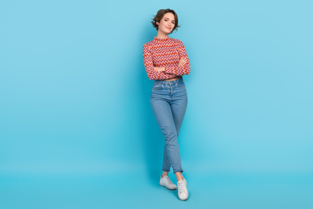 全尺寸的身体照片可爱自信的女士双臂交叉穿露脐衬衫牛仔裤运动鞋孤立的蓝色背景图片下载