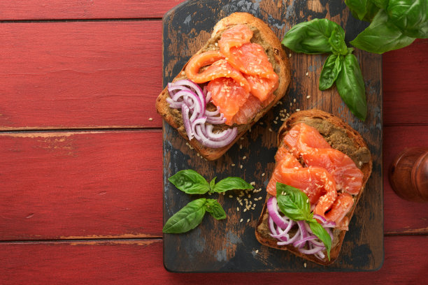 在旧的红色木制背景上，夹着咸鲑鱼、鳄梨鳄梨酱、红洋葱和罗勒的三明治或吐司。一套丹麦三明治。健康的食物，早餐。节食食物概念。前视图。图片下载