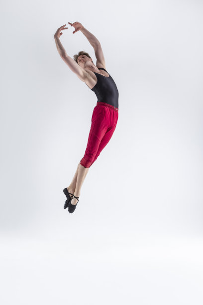 在白色的工作室里，年轻柔韧的运动员穿着红色紧身衣摆出飞翔的舞蹈姿势，双手举起并相连。图片下载