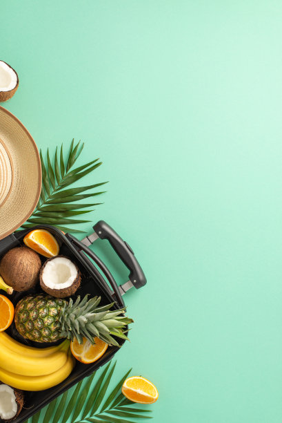 热带美学概念。上面的垂直视图照片，异国情调的水果在一个行李箱包围草帽和棕榈叶在孤立的蓝绿色背景与copyspace图片下载