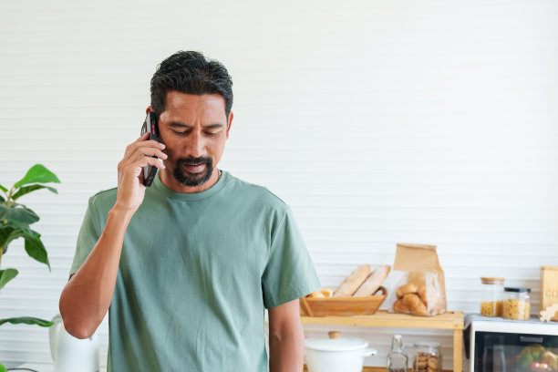 肖像，留胡子的亚洲男子在休闲日穿着t恤。在家里开始工作之前，站在厨房一边打电话一边等着做早餐。图片下载