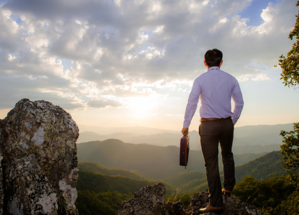 概念愿景，年轻的商人穿着舒适的休闲西装外套，手持商务包站在山顶，展望成功、竞争和领导者的理念。图片下载