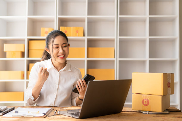小型创业公司，中小企业，企业家-快乐的亚洲女性使用智能手机或笔记本电脑接收和验证在线订单，准备盒子并从在线订单中发送包裹。图片下载