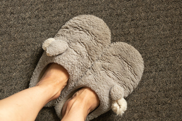 女脚和灰色柔软的室内拖鞋踩在灰色地毯上，室内鞋子的特写图片下载