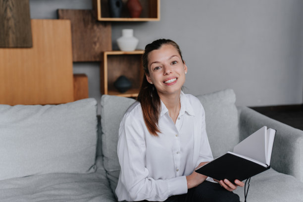 开朗的黑发美国女学生坐在家里舒适的沙发上开心地看着相机，手里拿着打开的书。办公室里穿白衬衫的白人女商人。图片下载