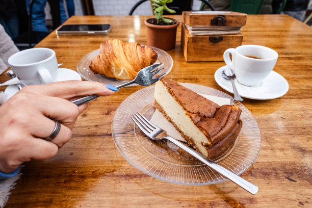 早餐。女人手里拿着叉子拿着一块芝士蛋糕放在一个玻璃板上，桌子上有一杯咖啡放在一个用金属勺子的盘子上，玻璃板上放着一个牛角面包和一杯茶。一切都在一张天然木桌上。美食。加那利群岛的特内里费岛图片下载