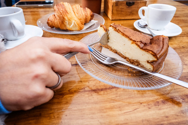 早餐。女人手里拿着叉子拿着一块芝士蛋糕放在一个玻璃板上，桌子上有一杯咖啡放在一个用金属勺子的盘子上，玻璃板上放着一个牛角面包和一杯茶。一切都在一张天然木桌上。美食。加那利群岛的特内里费岛图片下载