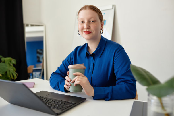 有创意的红发女人拿着咖啡杯在办公室的办公桌前对着镜头微笑图片下载