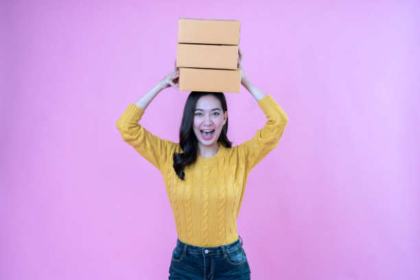 年轻的亚洲女子，长发，企业主头上顶着一个包裹箱，准备发送中小企业产品的订单，穿着黄色的外套在粉红色的现场摄影棚拍摄照片图片下载
