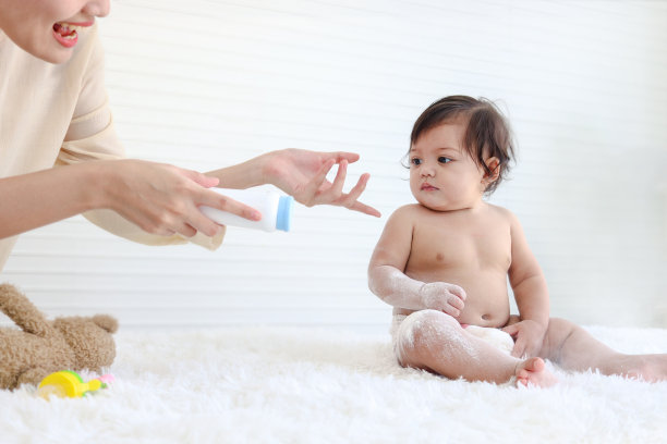 幸福微笑的蹒跚学步的女婴肖像与母亲手握粉扑瓶坐在蓬松的白色地毯上，母亲将滑石粉涂抹在小可爱的孩子女儿身上，童年照顾家庭图片下载