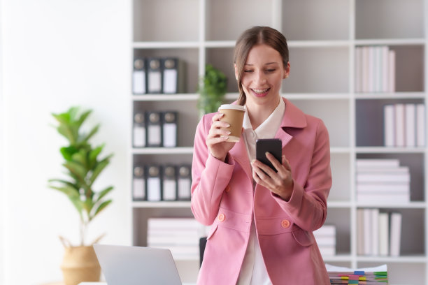 年轻漂亮的女商人一边拿着咖啡一边用智能手机。女人用手机，看电影，听音乐，网上购物，看直播。图片下载