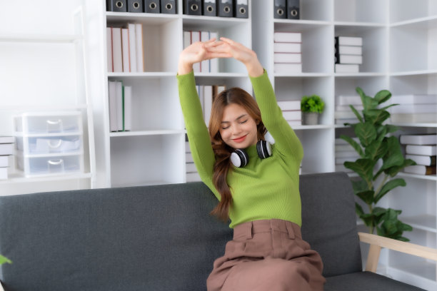 快乐的年轻女子坐在沙发上，在家里放松。她把双臂举过头顶。图片下载