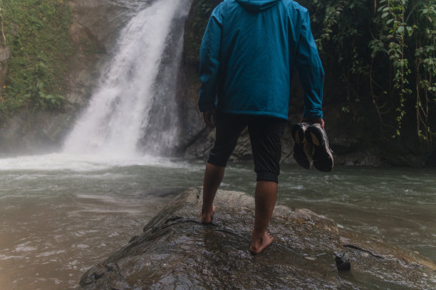 一名亚洲男子在热带雨林中拿着鞋子欣赏瀑布美景图片下载