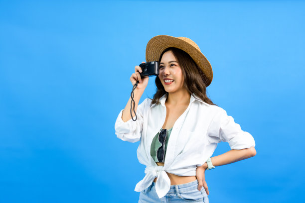 年轻的运动快乐的亚洲女人的肖像，黑色的长发和帽子微笑着用相机拍摄照片，在蓝色的工作室背景图片下载