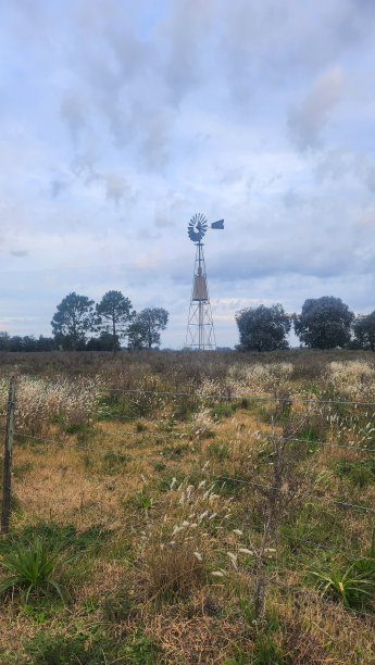 金属水磨在田野的中央，牧场和植被与树木在两侧和木材和铁丝栅栏在前景图片下载