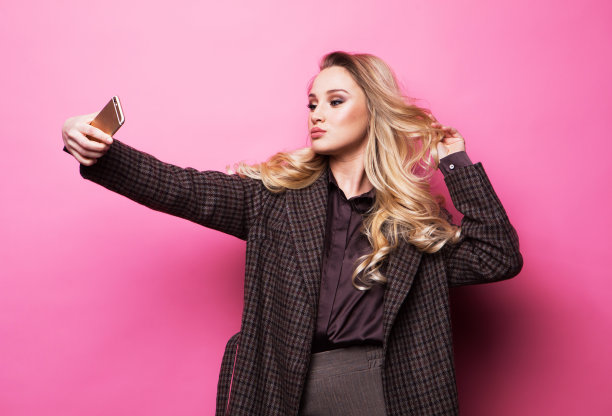 穿着大衣的金发女子在粉色背景下用智能手机自拍图片下载