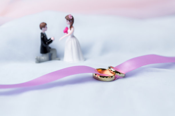 订婚的金结婚戒指。白色、粉色丝带背景的结婚金戒指。白玫瑰和红玫瑰上的戒指。结婚戒指是传统情侣婚礼的象征。图片下载