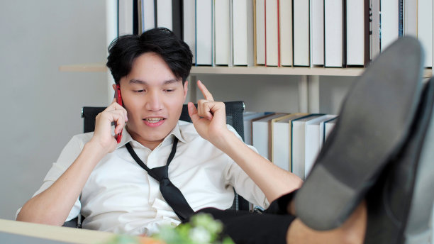 年轻的亚洲男子把脚放在办公室的桌子上，一边用智能手机聊天，一边休息。快乐商人手机图片下载