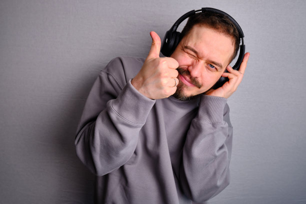 快乐的帅哥戴着耳机，穿着休闲运动衫，听着有声节目。在灯光工作室的背景下，很好的摆姿势。音乐概念与情感图片下载