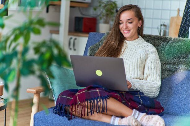 微笑着快乐可爱舒适的女人穿着白色温暖的毛衣，编织的袜子，盖着毯子放松地坐在沙发上用笔记本电脑在家里图片下载