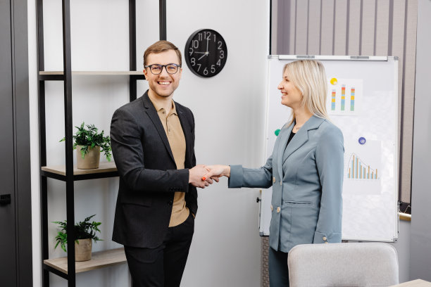 在办公室会议上，年轻女员工微笑着与男同事握手达成协议或成交。在简报会上，不同的商业伙伴愉快地握手打招呼。图片下载