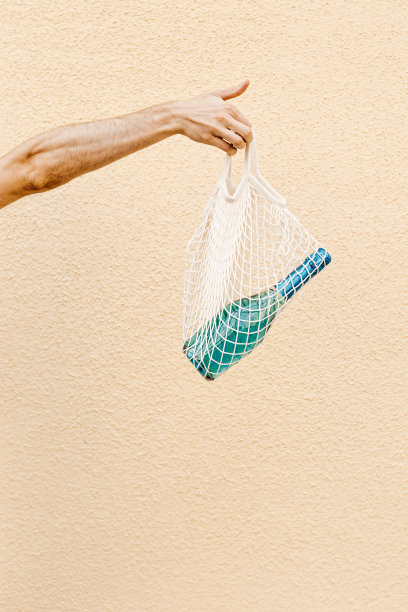 男士手里的网袋里装着蓝色的香槟起泡酒瓶，映衬着街道上米色的建筑墙壁，极简趋势的酒精饮料概念，夏天的生活方式都市照片，复制空间图片下载