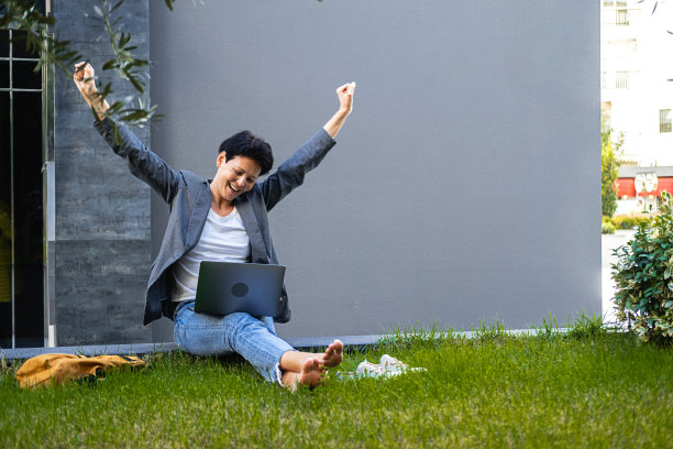 一个女人拿着笔记本电脑坐在草地上，高兴地举起双手。图片下载