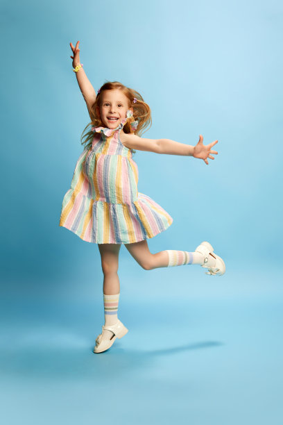 美丽的红发小女孩的全身肖像，孩子穿着可爱的条纹连衣裙，在蓝色的工作室背景下愉快地跳跃图片下载