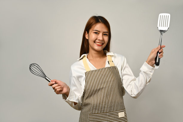 漂亮的亚洲家庭主妇系着围裙，拿着打蛋器和抹刀，放在白色的背景上准备做饭。图片下载