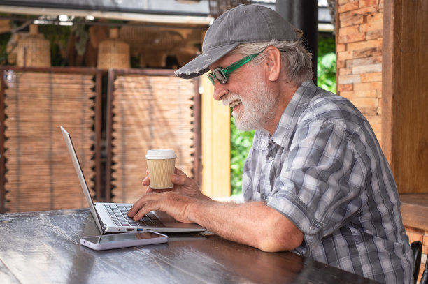 活跃的老人拿着咖啡杯，坐在户外的木桌上使用笔记本电脑。满脸笑容、蓄着胡子的老商人正在用无线技术上网图片下载